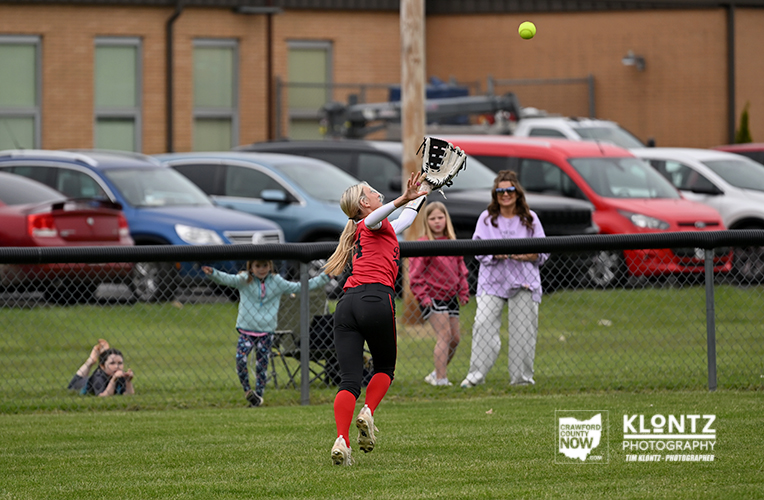 Young softball player in red reaches up to catch a high ball near the outfield fence, with onlookers and parked cars in the background.
