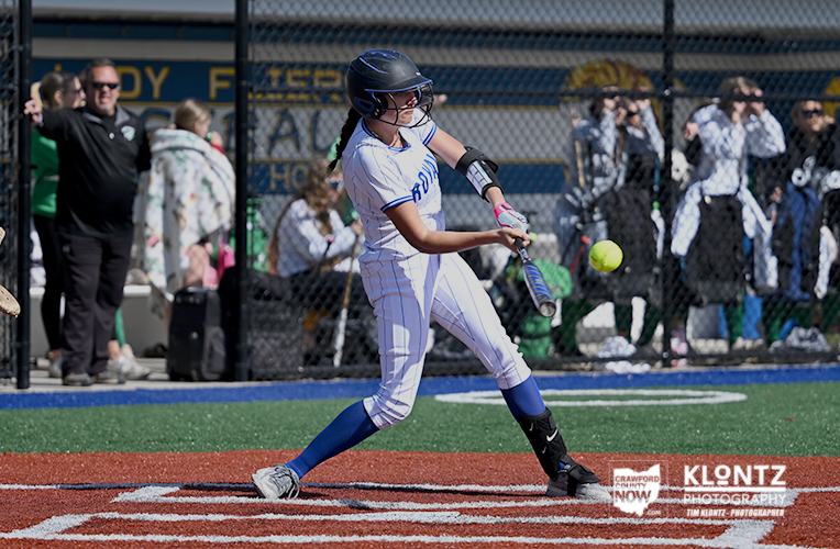 Female softball player in a white uniform and helmet swinging at a pitched ball during a game, with spectators in the fence-lined background.