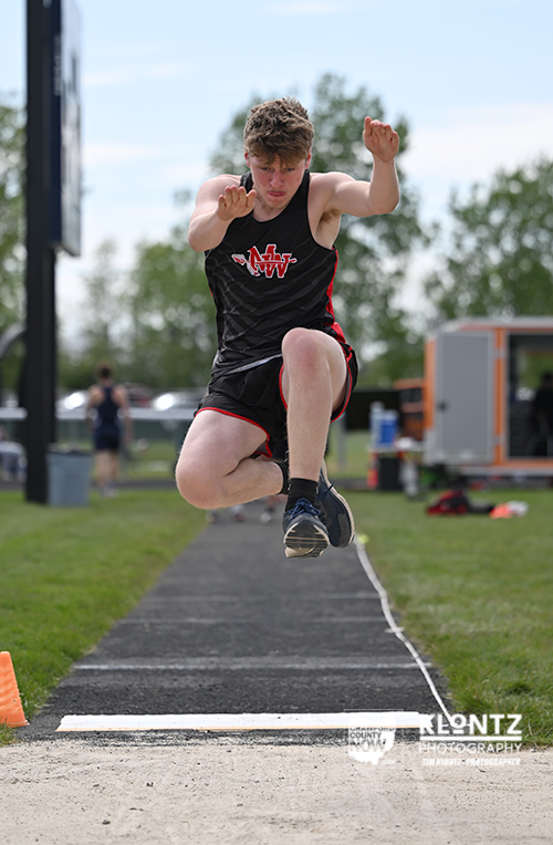 Male athlete in a black and red singlet and shorts mid-air during long jump on a track runway with a sand pit ahead and orange cones nearby.