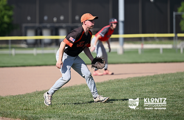 Young baseball infielder in black-and-orange uniform scoots to field a ground ball on a sunny field, glove ready, with a teammate in red in the background.