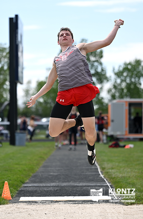 Male athlete in gray sleeveless top and red shorts leaps midair during a long jump on a track, sand pit ahead and spectators behind.