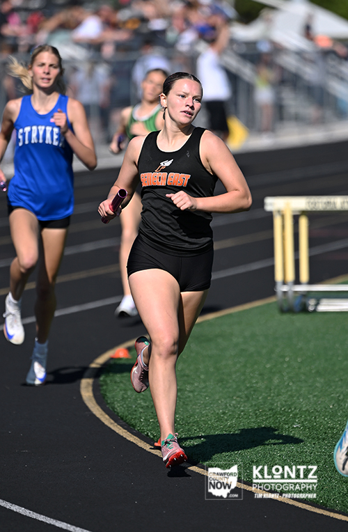 Female sprinter in a black uniform holding a baton, sprinting on a track with competitors behind her on the curve.