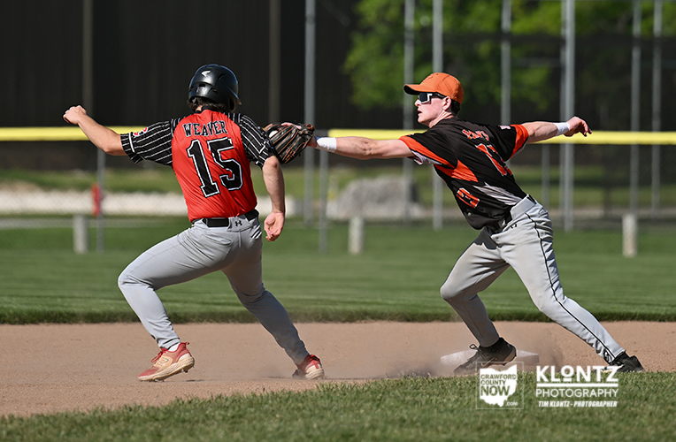 Runner in red #15 (Weaver) slides into a base as a fielder in black/orange reaches to tag him on a dirt infield.