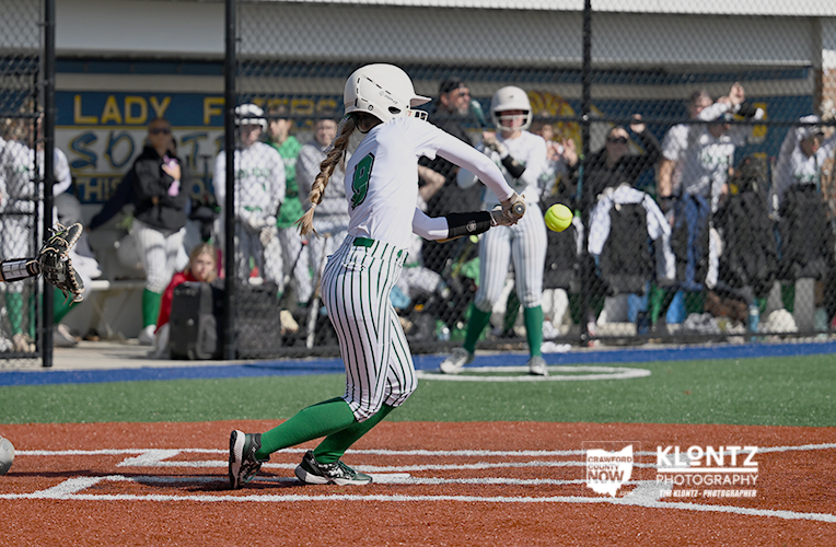 Female softball player in white uniform swings at a pitch at home plate, green socks visible, as spectators watch from behind a chain-link fence at the field.