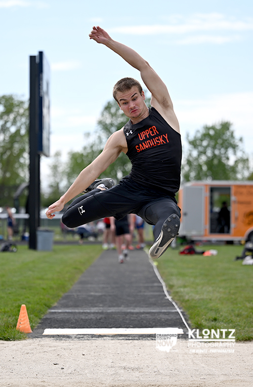 Male athlete in a black Upper Sandusky singlet jumps mid-air into a sand pit on a track runway, during a long jump event.