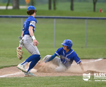 Baseball scene: a runner slides into a base as a fielder in blue watches from nearby, dust rising from the dirt.