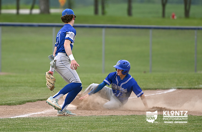 Baseball scene: a runner slides into a base as a fielder in blue watches from nearby, dust rising from the dirt.