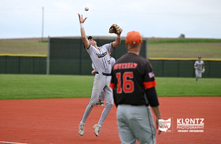 Baseball player in gray uniform leaps to catch a fly ball over the infield, while a teammate in a black jersey watches nearby.