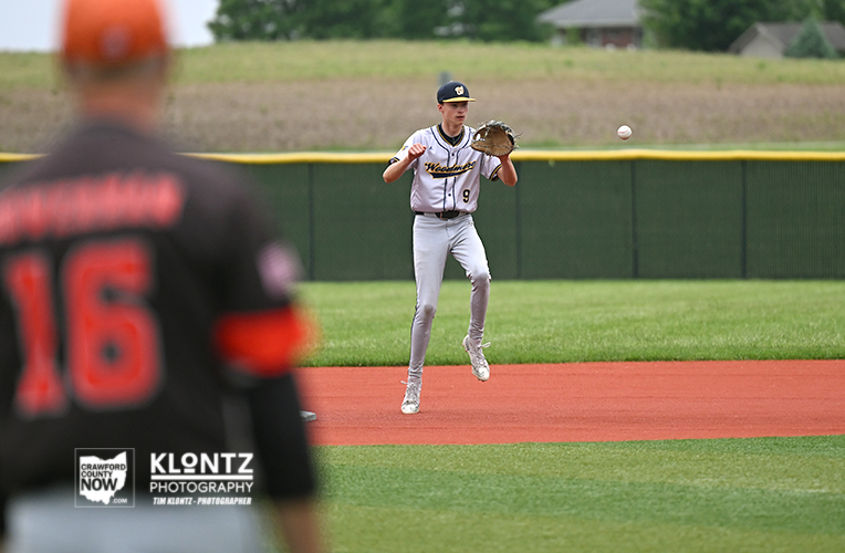 Baseball pitcher in gray uniform mid-pitch on a diamond, ball in the air, foreground player blurred in black and orange.