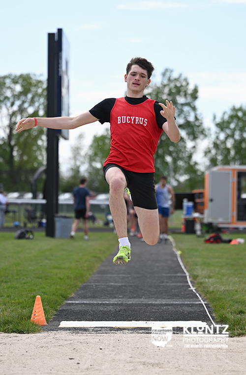 Young male athlete in a red Bucyrus jersey is mid-air during a long jump on a track, with the runway and sand pit in view.