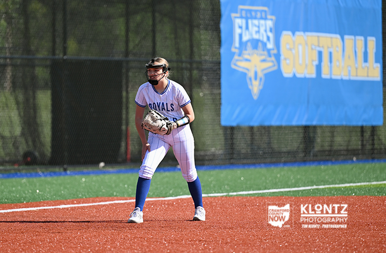 Softball player in a Royals uniform standing on the infield, glove ready for a ground ball, on a bright field with a blue softball banner in the background