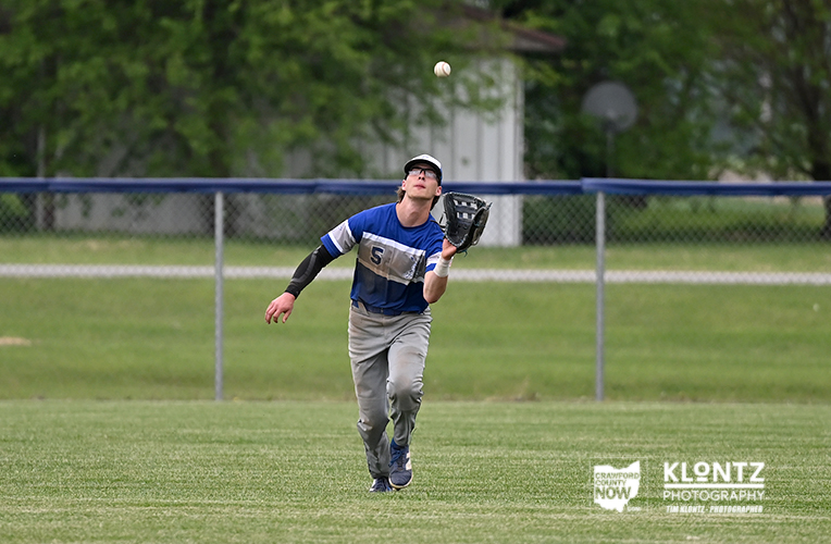 Baseball player in a blue jersey with number 5 runs toward a high ball, glove ready, on a grassy field with a chain-link fence behind him.