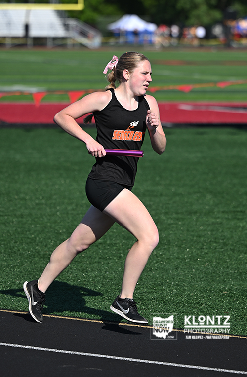 Young female relay runner in a black uniform sprints with a purple baton on an outdoor track, mid-stride.
