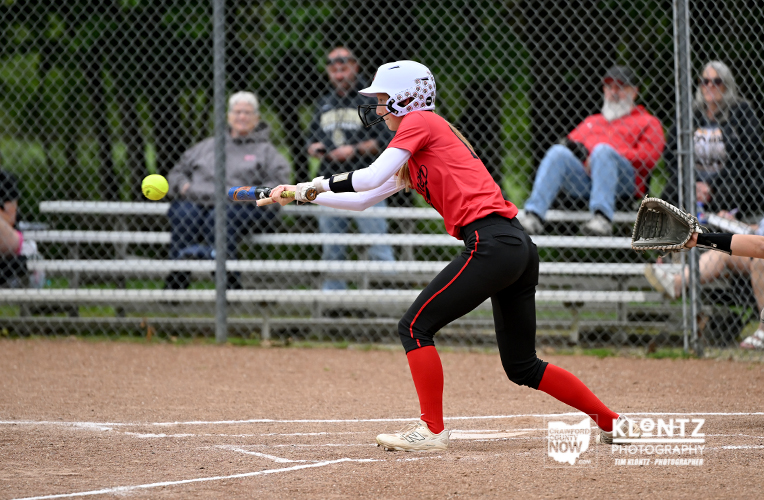 Female softball player in red uniform swings at a pitch during a game on a dirt field, with spectators behind a chain-link fence.