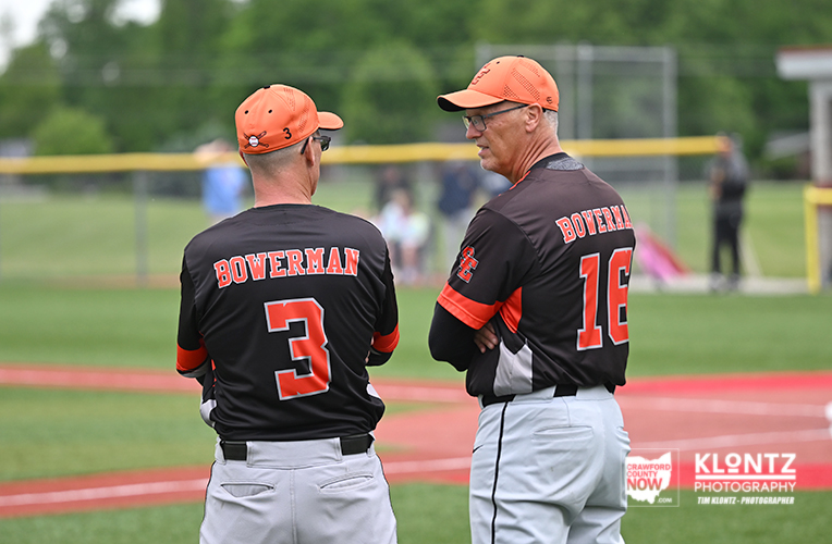 Two Bowerman baseball players stand on a green field with a net fence in the background, seen from behind in black-orange jerseys with numbers 3 and 16.