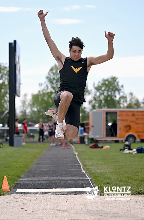 Male track and field athlete mid-jump during a long jump, arms raised, wearing a black singlet with a yellow emblem on a runway.