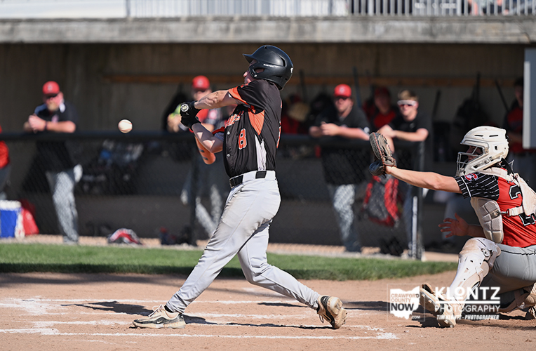 Baseball batter in black and orange swings at a pitch as the catcher in red gear reaches behind home plate behind him, with teammates watching in the dugout.