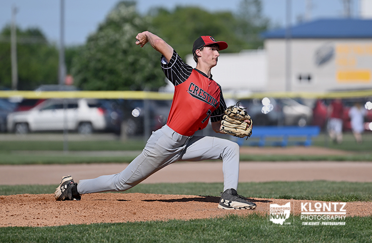 Baseball pitcher in a red Crestview uniform delivers a pitch from the mound on a sunny day.