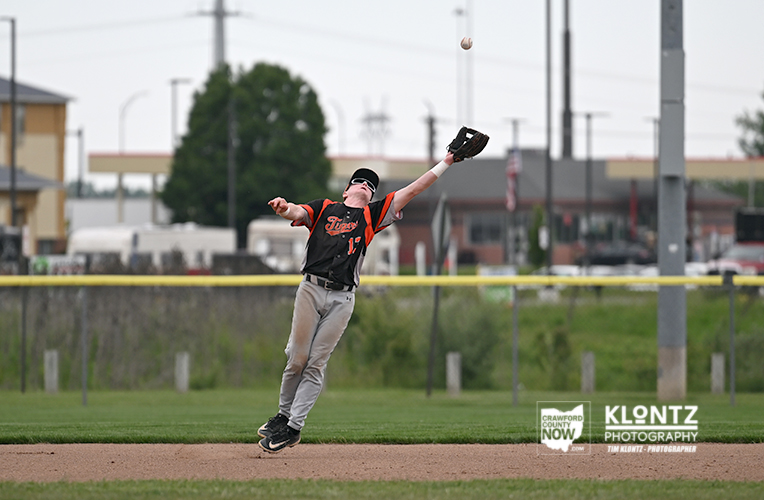 Baseball player in black-orange uniform leaps to catch a rising ball in the outfield grass with glove extended above his head on a sunny day