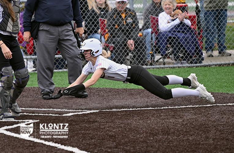 Female softball player in white uniform dives headfirst toward home plate, sliding on the infield as spectators watch.