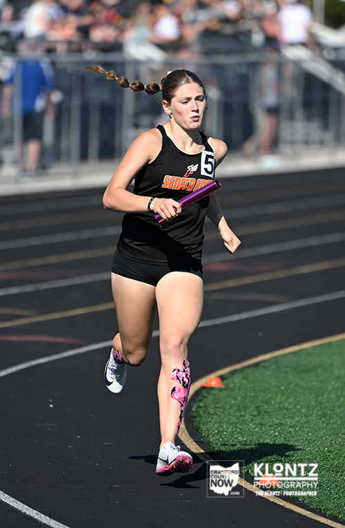 Female sprinter running on a track with a baton, wearing a black jersey and shorts, number 5 bib, braids trailing behind, crowd in the stands in the background.