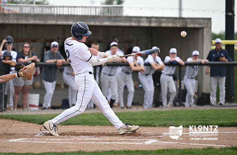 Baseball batter in white uniform swings at a pitched ball at home plate with teammates and spectators watching from the dugout in the background.
