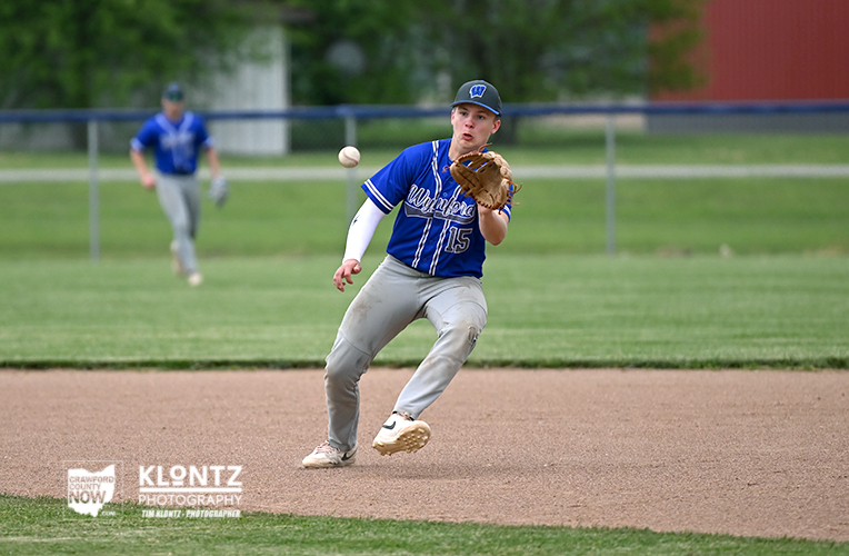 Baseball infielder in a blue uniform (number 15) crouches to field a ground ball on the infield dirt, with a teammate in blue in the background.
