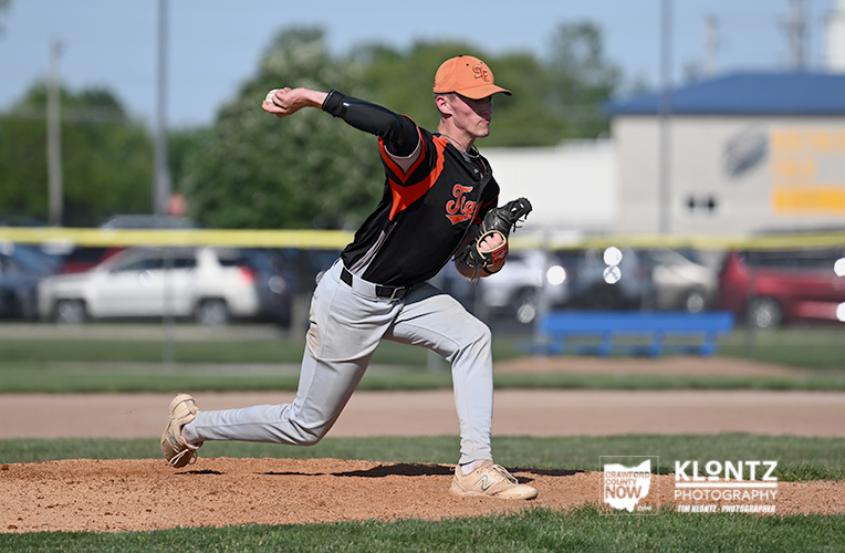 Baseball pitcher in an orange and black uniform throwing a pitch on the mound, glove up and back foot off the dirt, with a parking lot and field in the background.