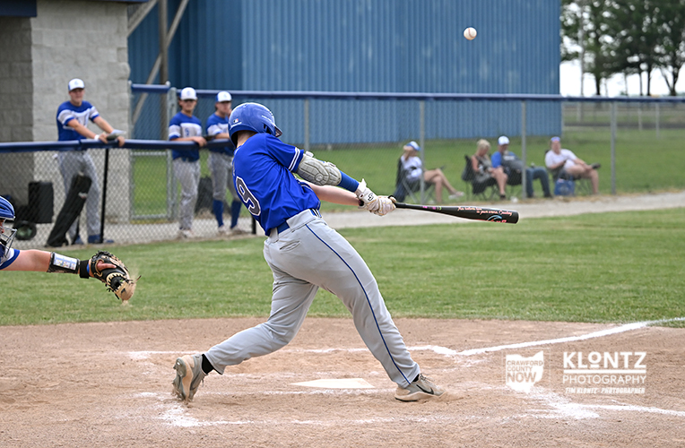 Baseball batter in blue uniform swinging at a pitched ball during a game, with a catcher and teammates in the dugout behind him and spectators in the stands nearby.