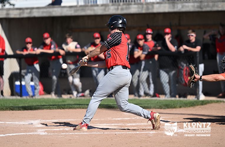 Baseball player in a red and black uniform mid-swing at home plate with teammates watching in the Dugout area behind the fence.