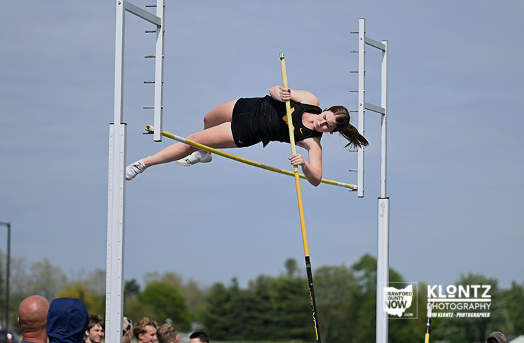 Female athlete in a black outfit vaults over a high bar with a yellow pole during an outdoor pole vault competition.