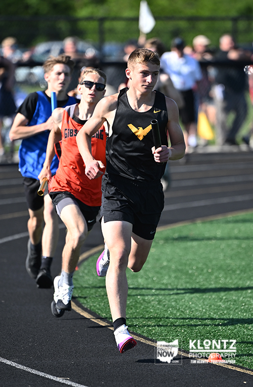 Three male relay runners sprint along a track; the lead runner in a black uniform with a yellow logo holds a baton, with two followers closely behind on a sunny day.