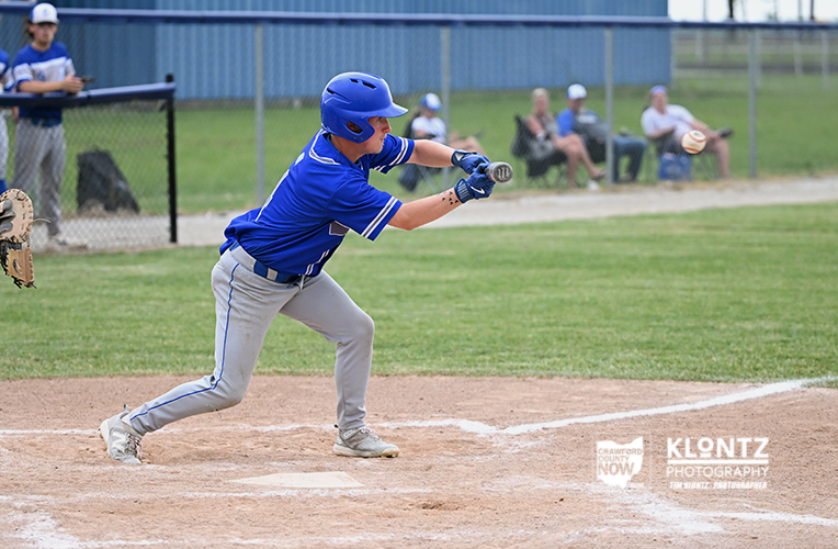 Youth baseball player in a blue uniform and helmet swings at a pitch on a dirt infield, with spectators watching from the outfield fence.
