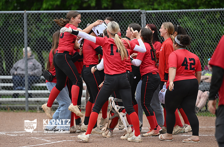 Youth girls' softball team in red uniforms celebrate together on the infield after a game, near a chain-link fence.