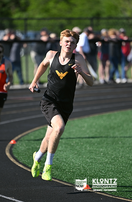 Young male relay runner in a black uniform with a yellow emblem sprints on an outdoor track, baton in hand, with spectators in the background.