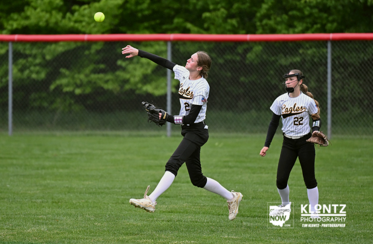Two female softball players in white and black uniforms play on a grassy field; one pitcher throws the ball while a fielder watches from behind near a chain-link fence.
