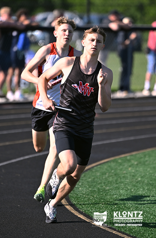 Two male relay runners sprint on a track; the front runner in a black uniform with a red emblem holds a baton as the second racer in red and white closes in, with spectators in the background.