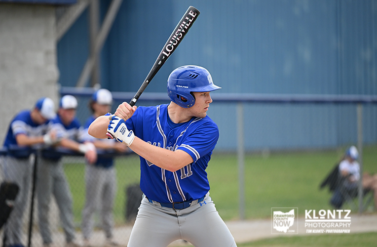 Baseball batter in blue uniform and helmet, gripping a Louisville Slugger bat, ready to swing; teammates in blue sit in the blurred dugout in the background.