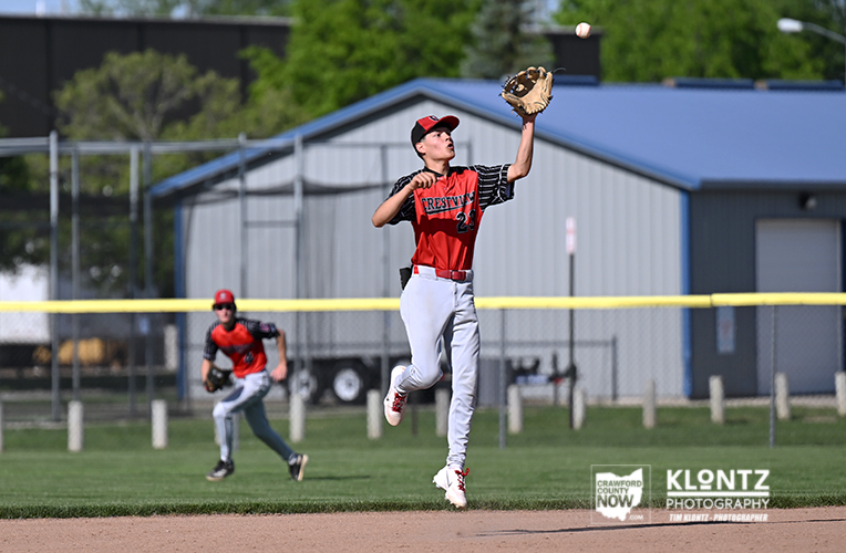 Young baseball pitcher in red-and-black uniform leaps with glove raised to catch a ball during a game on a sunny field; another fielder watches in the background.
