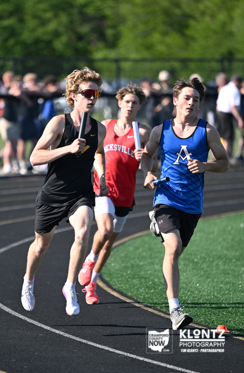 Three male runners relay race on an outdoor track; foreground runner in a blue jersey with an 'M' leads, two others in black and red behind, each holding batons.