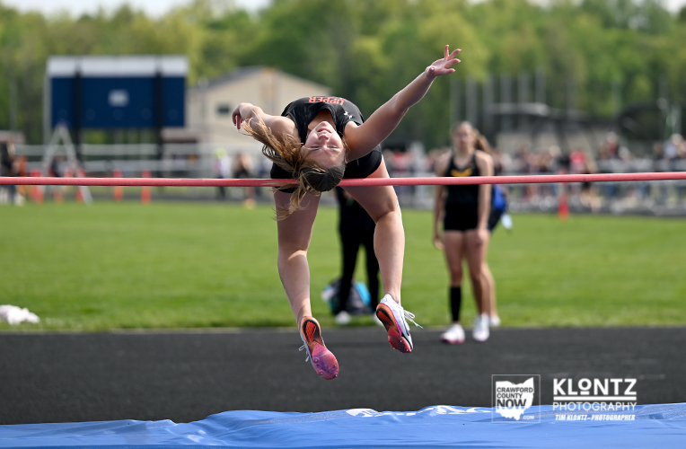 Female athlete clears the high jump bar in a track and field competition, midair over the bar on a sunny day.