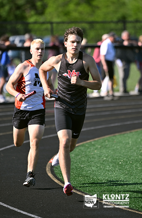 Two boys in a relay race sprint on an outdoor track, each holding a baton as spectators watch in the background.