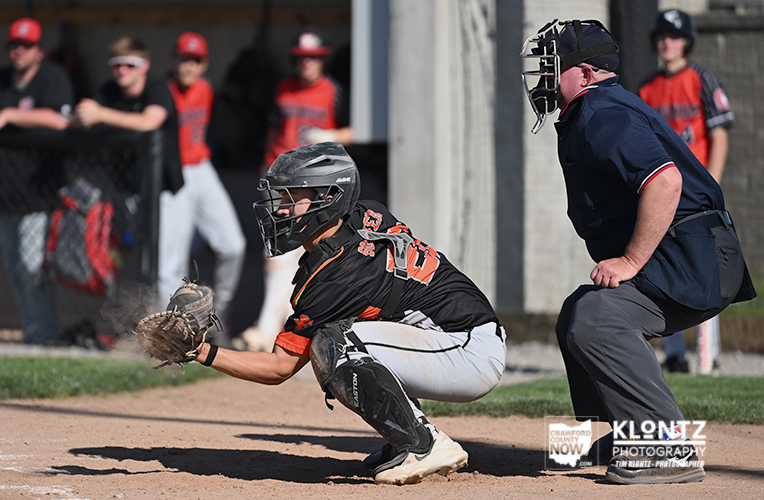 Baseball catcher in black and orange gear slides toward home plate with glove ready as an umpire watches nearby.