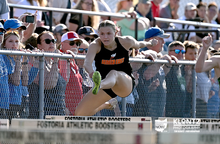 Female hurdler in a black Seneca East uniform leaps over a track hurdle as spectators watch from the stands behind a chain-link fence.