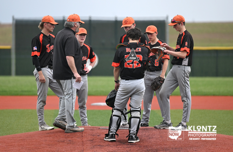 Youth baseball team huddles on the pitcher's mound as a coach speaks to them on a sunny field with a red clay infield.