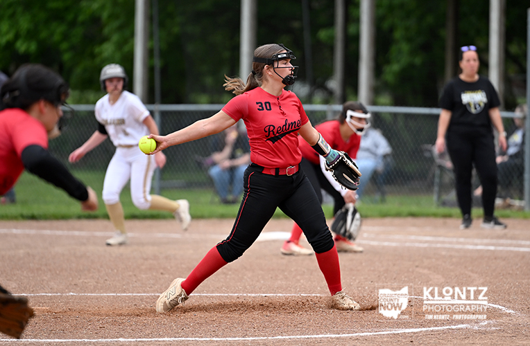 Female softball pitcher in a red jersey (number 30) throws a pitch from the mound on a dirt field, wearing a protective mask and red socks.