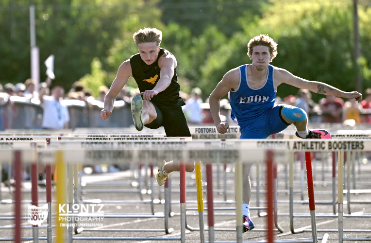 Two male athletes sprint over hurdles in an outdoor track race, crowd watching in the background, one in black and one in blue.