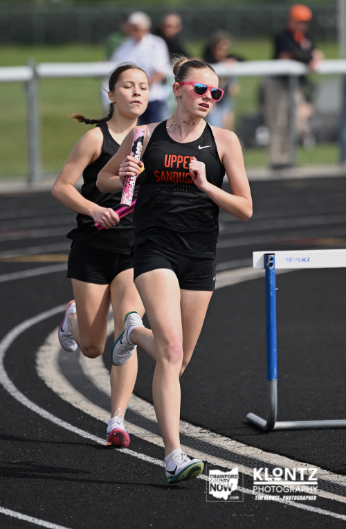 Two female sprinters race on a track, one in front wearing sunglasses and a black Nike singlet, the other closely behind with a pink baton-like object; both in mid-stride on a curved track with spectators in the background.