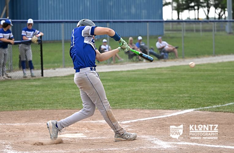 Baseball player in blue uniform swings a bat at a pitched ball during a game on a dirt field, with teammates watching from the dugout in the background.