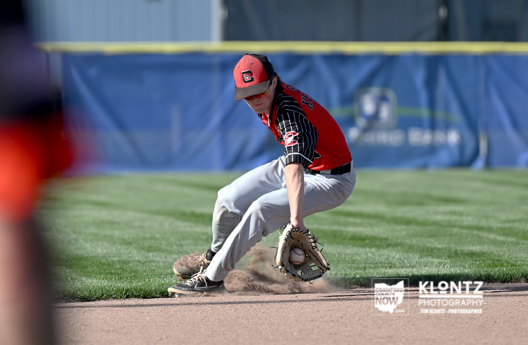 Baseball infielder in red and black uniform slides to scoop a grounder with a glove near the infield dirt.